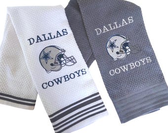 Op de afbeelding: Twee Dallas Cowboys handdoeken. Eén is wit met een donkergrijze streep en de andere is grijs. Beide handdoeken hebben de woorden "DALLAS COWBOYS" in wit geborduurd, samen met een voetbalhelm.