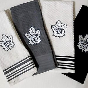 Puede incluir: Cuatro paños de cocina en blanco, gris y negro, cada uno con el logotipo de los Toronto Maple Leafs. Los paños tienen un patrón texturizado y un borde a rayas. El logotipo es una hoja de arce estilizada con el nombre del equipo.