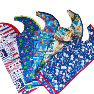 May include: Five colorful bibs with different patterns, including sailboats, flamingos, and dogs. The bibs are made of fabric and have a red, blue, and white color scheme.