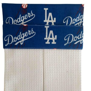 May include: A white waffle-weave kitchen towel with a blue top band featuring the Los Angeles Dodgers logo and the word "Dodgers" in white script. The top band also displays the team's "LA" logo.