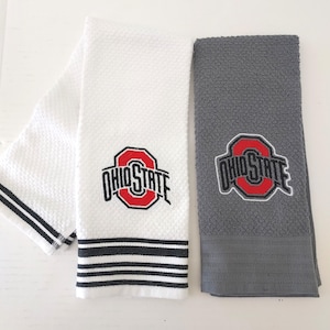 May include: Two rectangular kitchen towels, one white with black stripes and one solid grey. Both feature the embroidered Ohio State logo in red, black, and white. The white towel has a textured waffle weave, while the grey towel has a solid finish.