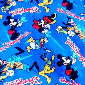 Puede incluir: Tela azul con un patrón repetido de personajes de Disney, incluyendo Mickey Mouse, Minnie Mouse, Donald Duck y Pluto. Las palabras "Walt Disney World" y "Disneyland Resort" también son visibles en texto rojo y blanco.