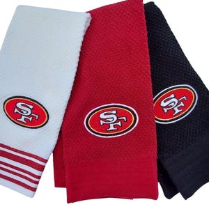 Peut inclure: Trois serviettes pliées, blanches, rouges et noires. Chaque serviette présente un logo brodé des 49ers de San Francisco. La serviette blanche a des rayures rouges en bas. Idéal pour les fans de sport.