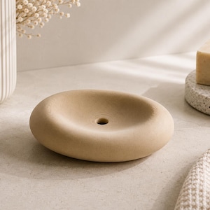Könnte beinhalten: Eine runde, beige Seifenschale mit glatter, matter Oberfläche und einem einzelnen Abflussloch in der Mitte. Die Schale hat eine leicht konkave, schalenartige Form mit dicken, abgerundeten Kanten, die eine stabile Basis für Seifenstücke bietet.