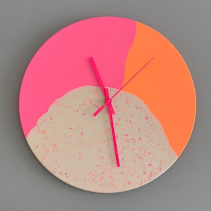 Könnte beinhalten: Eine runde Wanduhr mit einem Color-Blocking-Design in Neonpink und leuchtendem Orange. Der untere Teil besteht aus einer beigen Oberfläche im Terrazzo-Stil mit kleinen rosa Sprenkeln. Die Uhr hat drei dünne, neonpinke Zeiger.