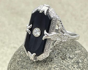 May include: A silver ring featuring a rectangular black onyx stone with a central, round, clear gemstone. The ring has an ornate, filigree design with floral and leaf accents. The ring is set on a gray stone.