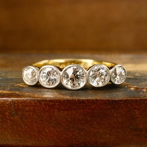 May include: A gold band ring featuring five round, bezel-set diamonds arranged in a horizontal row. The center diamond is the largest, with two smaller diamonds on each side, all set in a polished metal frame that highlights the stones.