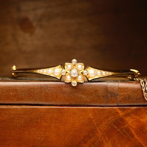 May include: A gold-toned bracelet featuring a floral design at its center. The flower is composed of pearls and a central diamond, with additional diamonds and pearls along the bracelet's curved arms. The bracelet is resting on a wooden surface.