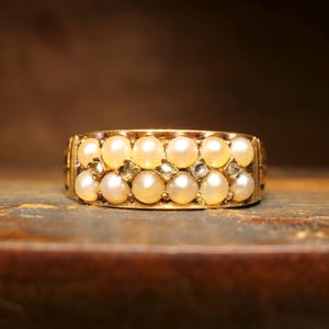 Könnte beinhalten: Ein Goldring mit zwei horizontalen Reihen kleiner, runder, cremefarbener Perlen. Zwischen den beiden Perlenreihen sind mehrere kleine, klare, facettierte Steine in das Metallband eingefasst, was ein dekoratives, mehrschichtiges Design erzeugt.