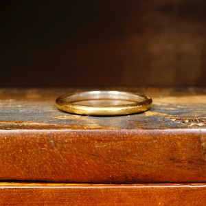 May include: A gold-colored ring with a textured surface. The ring is set on a wooden surface with a dark background. The ring appears to be a wedding band or a similar type of jewelry.