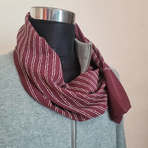 May include: A burgundy scarf with a white vertical stripe pattern is draped around a mannequin's neck. The scarf is paired with a gray zip-up sweater. The scarf's fabric has a smooth texture, and the overall look is stylish and warm.