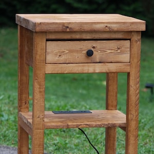 May include: A rustic wooden nightstand with a single drawer and two shelves. The nightstand is made of light brown wood and has a black knob on the drawer. A black power cord is visible.