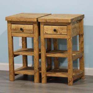 May include: Two rustic wooden nightstands. Each nightstand has a drawer with a black knob and two open shelves. The nightstands are made of light brown wood and have a simple, sturdy design.