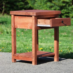 May include: A wooden end table with a single drawer that is open. The table is stained a dark reddish brown color and has a simple design. The table is sitting on a gray asphalt surface.