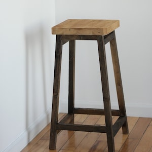 May include: A wooden stool with a dark brown stained frame and a natural wood top. The stool is standing on a wooden floor.