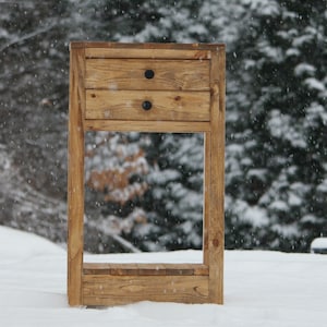 May include: A wooden nightstand with two drawers and a shelf. The nightstand is painted a light brown color and has black metal drawer pulls. The nightstand is sitting in a snowy landscape.