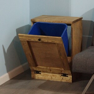 May include: A wooden trash can with a blue bin inside and a hinged door. The trash can is sitting on a wooden floor.
