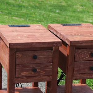 May include: Two wooden end tables with two drawers each. The tables have a dark reddish-brown stain and black metal drawer pulls. Each table has a built-in power outlet on the top surface.