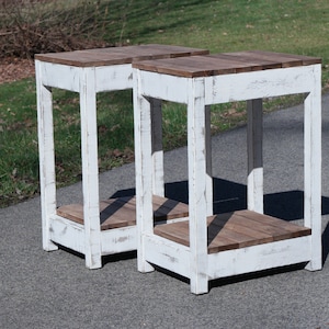 May include: Two white painted wooden end tables with a distressed finish. The tables have a square top and a single shelf. The tables are sitting on a gray asphalt driveway.