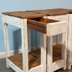 May include: Set of three rustic wooden side tables. Each table features a distressed white painted frame with a natural wood top and drawer. The drawer is pulled out on one table, revealing the interior.