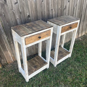 May include: Two white painted wooden end tables with a drawer and a shelf. The tables have a distressed wood top.