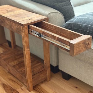 May include: A wooden side table with a drawer, stained in a warm brown hue. The drawer is partially extended, revealing its interior and metal drawer slides. The table has a lower shelf and is positioned next to a sofa.