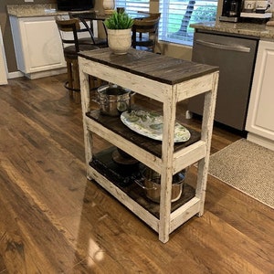 May include: White distressed wood kitchen cart with three shelves. The cart has a dark wood top and is filled with kitchen items, including pots, pans, and a plate.