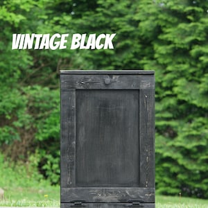 May include: A black wooden trash can with a single door and two metal handles. The can is sitting on a gray asphalt surface. The text "Vintage Black" is visible in the image.