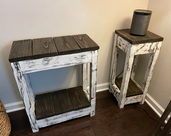 May include: Two whitewashed wooden end tables with dark brown wooden tops and shelves. The tables have a distressed look with white paint chipping off to reveal the natural wood underneath.