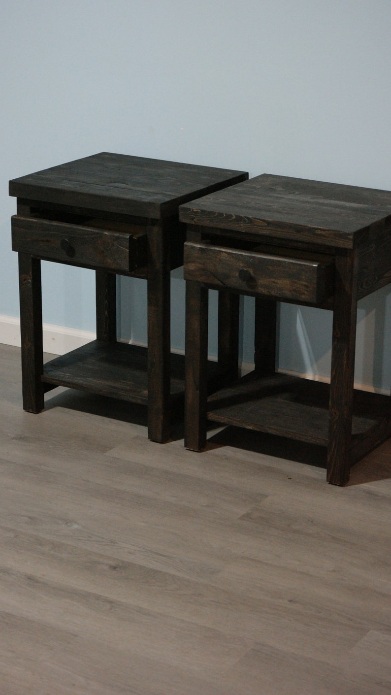 May include: A pair of dark brown wooden nightstands, each featuring a square top, a single pull-out drawer with a small round knob, four straight legs, and an open lower shelf. The wood grain is visible throughout the dark stained finish.