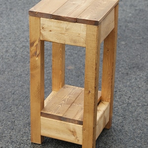 May include: A wooden two-tiered stand with a natural finish. The stand has a square top and bottom shelf, and is made of solid wood planks.