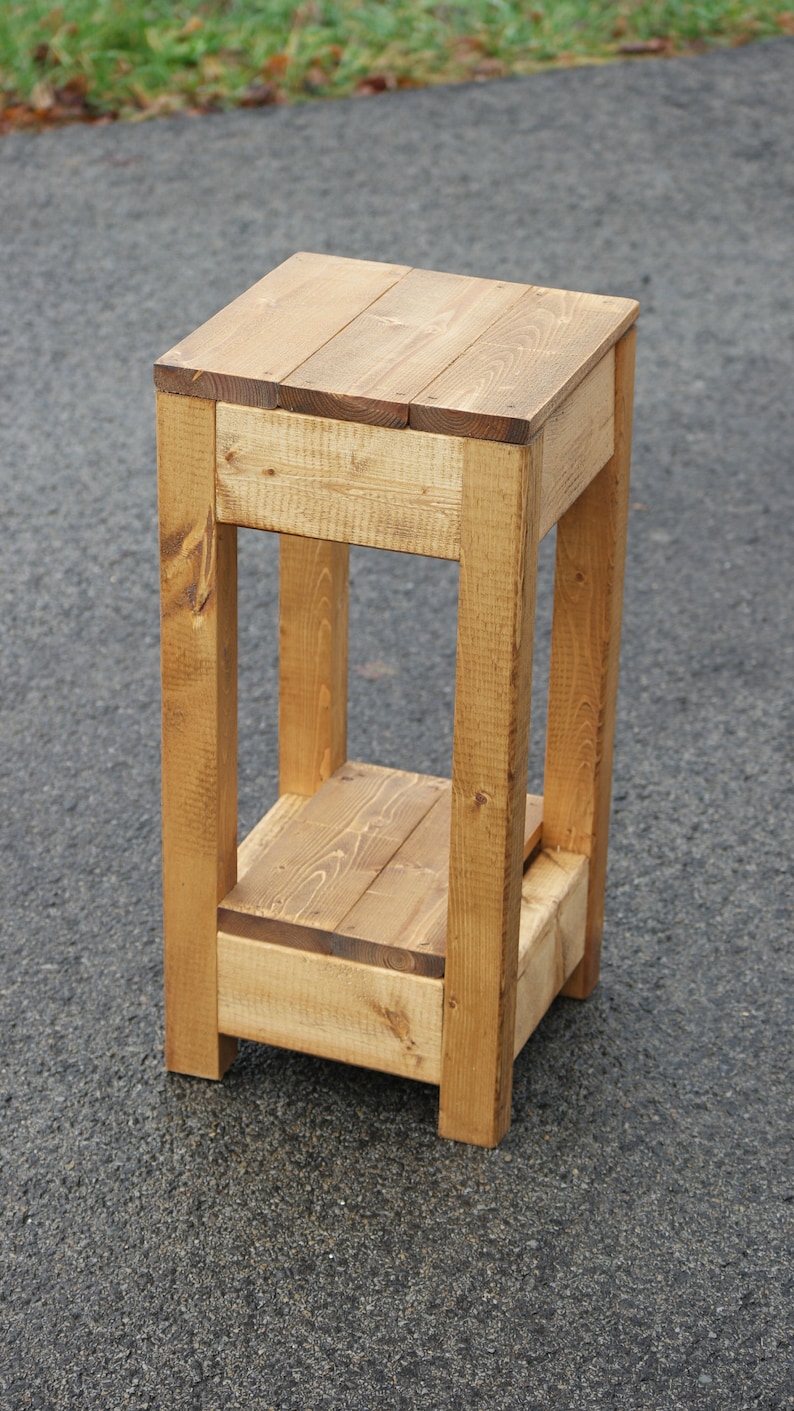 May include: A wooden two-tiered stand with a natural finish. The stand has a square top and bottom shelf, and is made of solid wood planks.