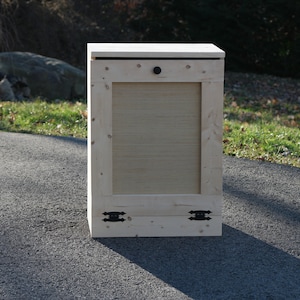 May include: A wooden storage bin with a hinged lid and a small black knob. The bin has two black metal hinges on the bottom and is sitting on a paved driveway.