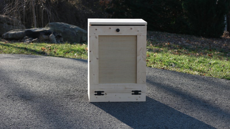May include: A wooden storage bin with a hinged lid and a small black knob. The bin has two black metal hinges on the bottom and is sitting on a paved driveway.