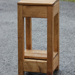 May include: A rustic wooden shelf unit with two shelves. The shelf is made of natural wood with a light brown finish. The shelf is standing on a gray asphalt surface.