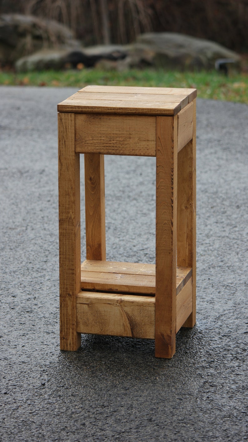 May include: A rustic wooden shelf unit with two shelves. The shelf is made of natural wood with a light brown finish. The shelf is standing on a gray asphalt surface.