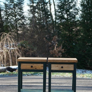 May include: Two matching wooden nightstands featuring a natural light wood top and drawer front contrasted by a dark green painted frame. Each piece includes a single drawer with a small round black knob and an open lower shelf for storage.