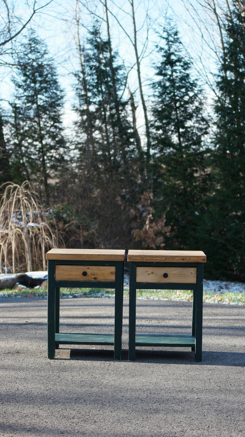 May include: Two matching wooden nightstands featuring a natural light wood top and drawer front contrasted by a dark green painted frame. Each piece includes a single drawer with a small round black knob and an open lower shelf for storage.