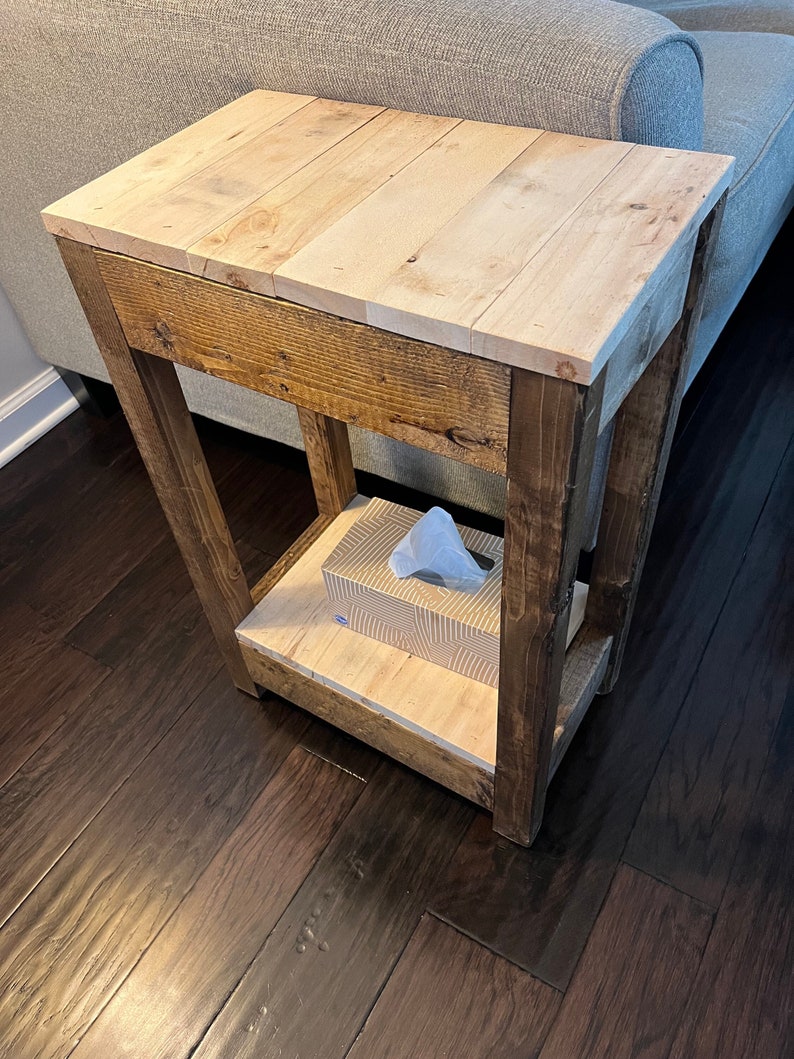 May include: A rustic wooden end table with a natural wood top and a lower shelf. The table has a distressed brown finish and is made of reclaimed wood. A box of tissues sits on the lower shelf.