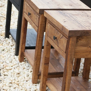 May include: Three wooden side tables with a rectangular top, a single drawer featuring a small round knob, and a lower shelf. Two tables are finished in a medium brown wood stain, while the third table in the back is finished in black.