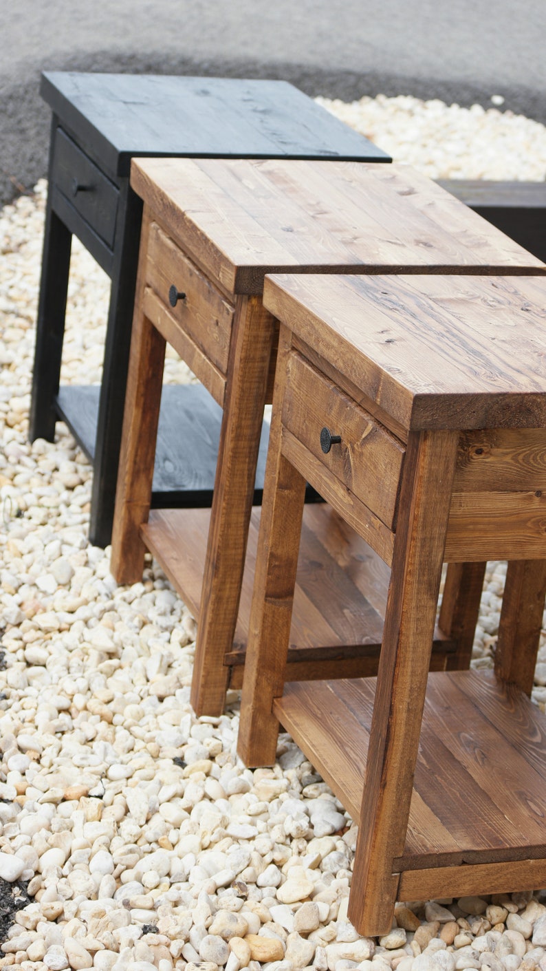 May include: Three wooden side tables with a rectangular top, a single drawer featuring a small round knob, and a lower shelf. Two tables are finished in a medium brown wood stain, while the third table in the back is finished in black.