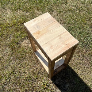 May include: A small wooden end table with a single shelf. The table is made of natural wood with a light brown finish. The table is sitting on a patch of green grass.