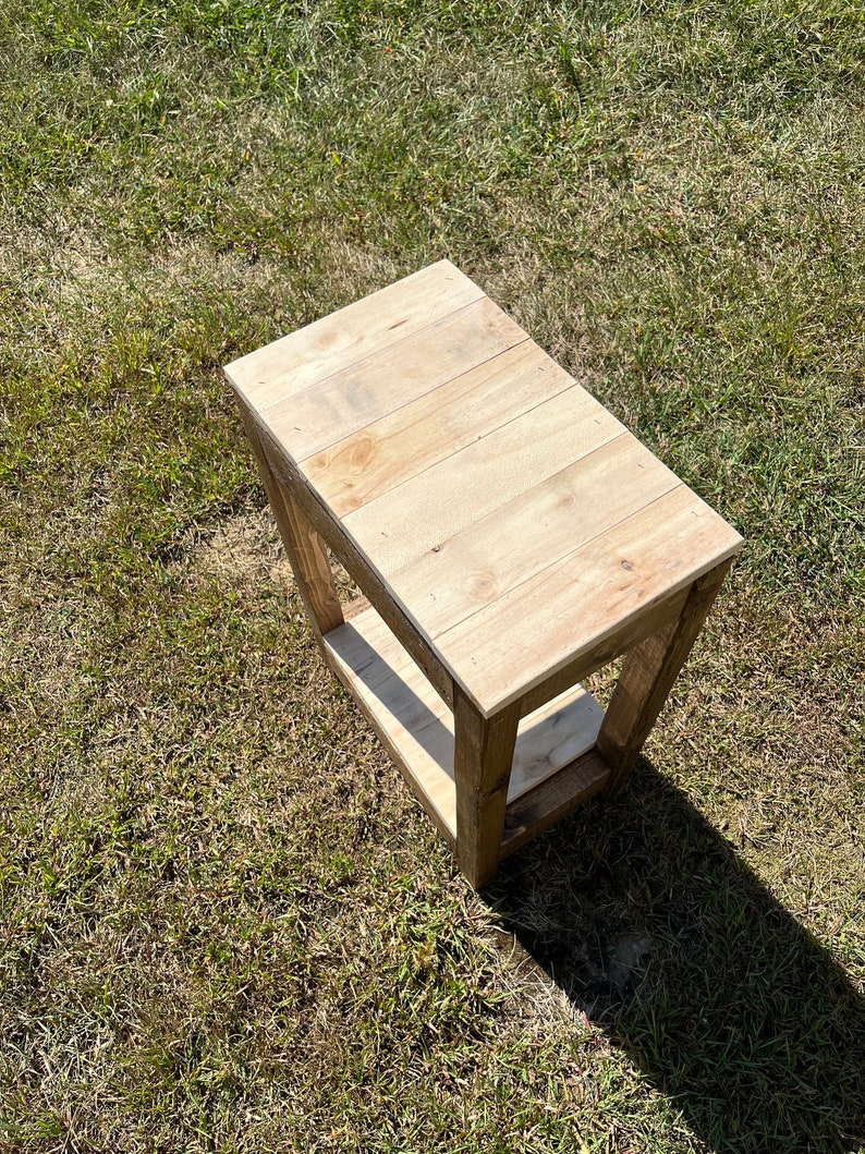 May include: A small wooden end table with a single shelf. The table is made of natural wood with a light brown finish. The table is sitting on a patch of green grass.
