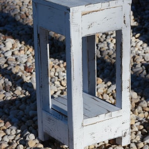 May include: White wooden stool with a distressed finish and a shelf. The stool is sitting on a bed of small white rocks.