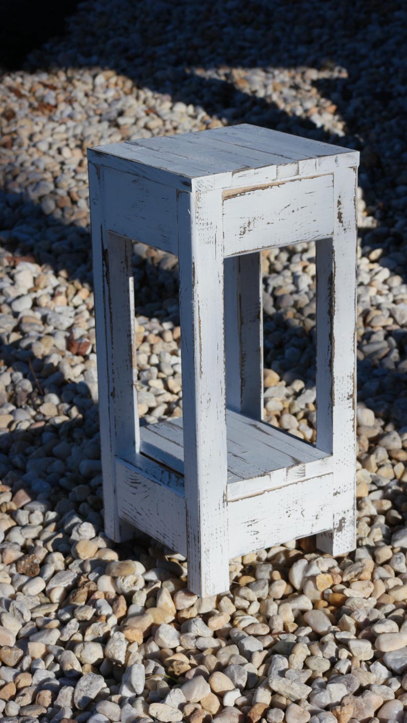 May include: White wooden stool with a distressed finish and a shelf. The stool is sitting on a bed of small white rocks.