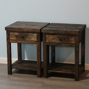May include: A pair of matching dark brown wooden nightstands, each featuring a single pull-out drawer with a small dark knob and an open lower shelf. The tables have a rectangular top surface and four straight legs, showcasing a visible wood grain.