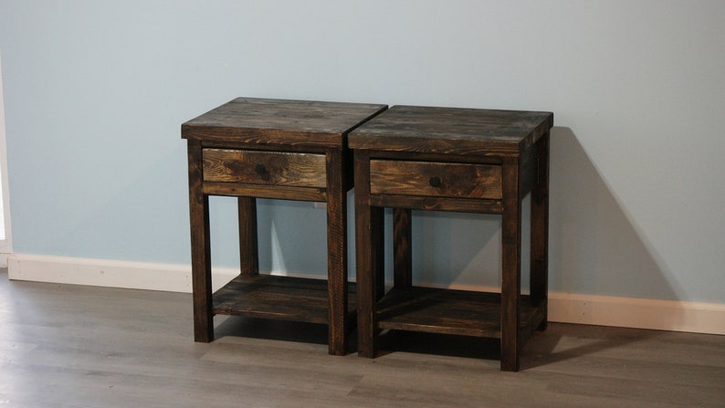 May include: A pair of matching dark brown wooden nightstands, each featuring a single pull-out drawer with a small dark knob and an open lower shelf. The tables have a rectangular top surface and four straight legs, showcasing a visible wood grain.