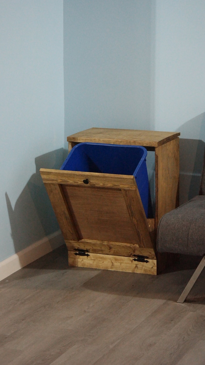 May include: A wooden trash can with a blue bin inside and a hinged door. The trash can is sitting on a wooden floor.