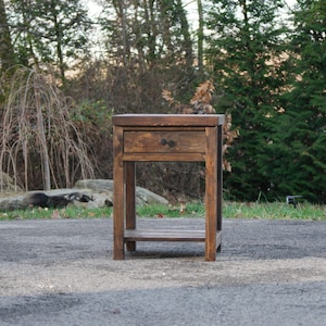 May include: A dark brown wooden nightstand featuring a single drawer with a small round knob and an open lower shelf. The piece is constructed with four straight legs and has a visible wood grain texture throughout its rectangular frame.