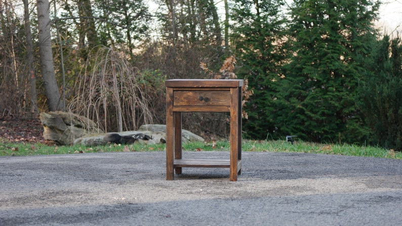 May include: A dark brown wooden nightstand featuring a single drawer with a small round knob and an open lower shelf. The piece is constructed with four straight legs and has a visible wood grain texture throughout its rectangular frame.